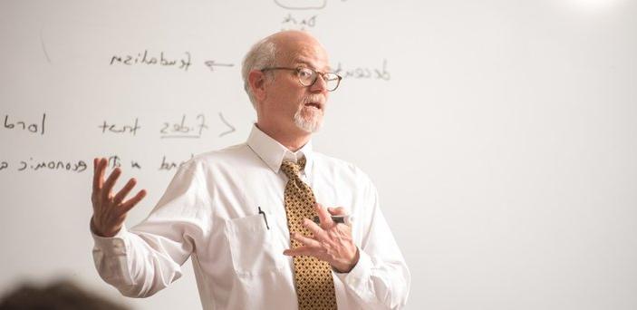 A professor teaching in front of a white board with words on it including Dark Ages, urbanism, feudalism and lord.